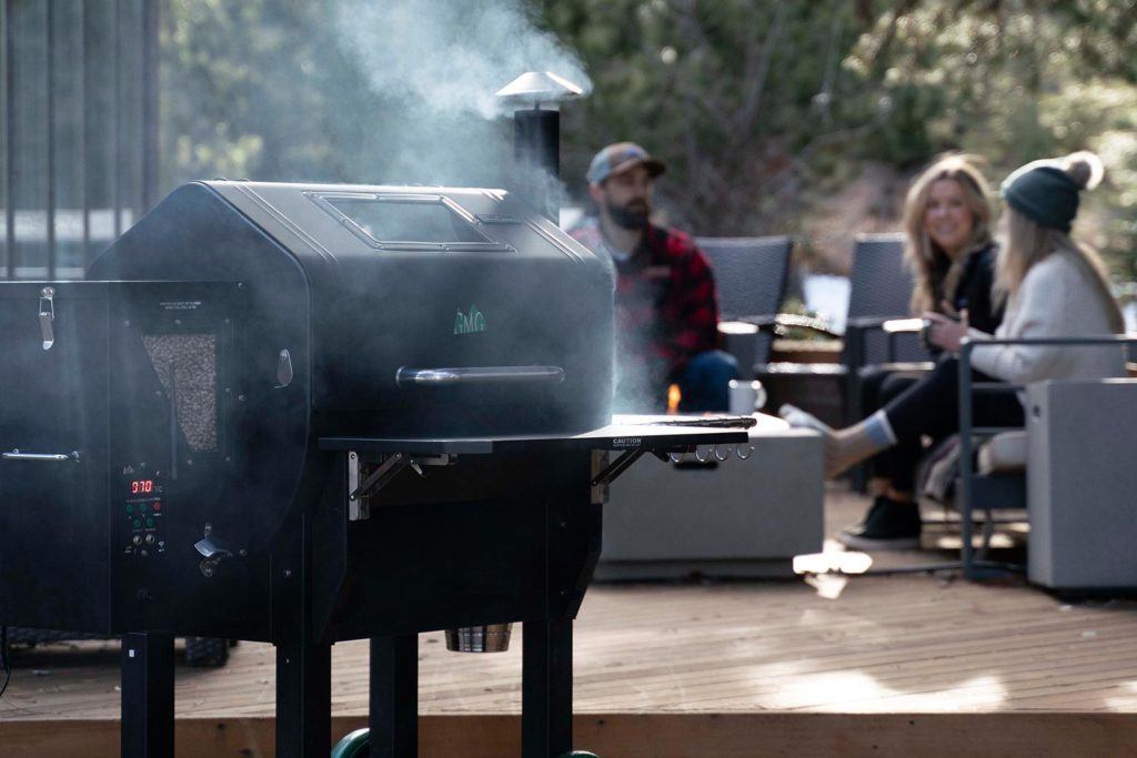 a smoker BBQ with wood pellet attachment smoking outdoors while people socialize on a deck in the background