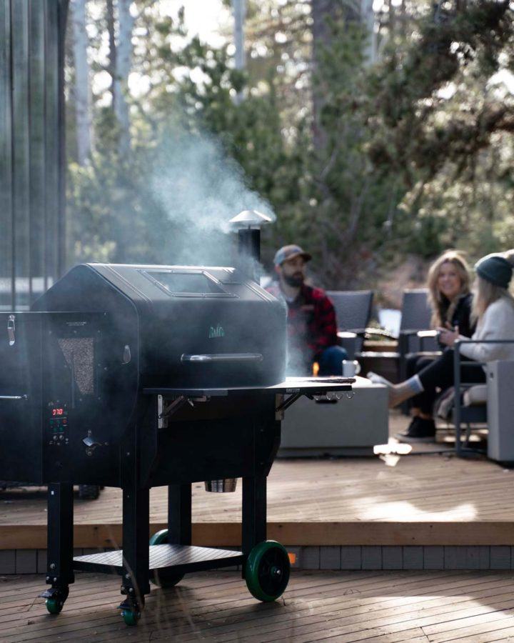 a smoker BBQ with wood pellet attachment smoking outdoors while people socialize on a deck in the background
